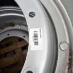 Close-up of a silver car wheel rim with a circular hole and a white barcode label. A second bolt hole is visible nearby, with dirt and wear on the rim edge.