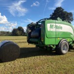 Green McHale baler in a field with a large wrapped round bale on the left and a tractor beside it under a blue sky.