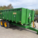 Green farm trailer with large yellow wheels, hitched to a tractor, parked on a gravel lot with trees in the background.
