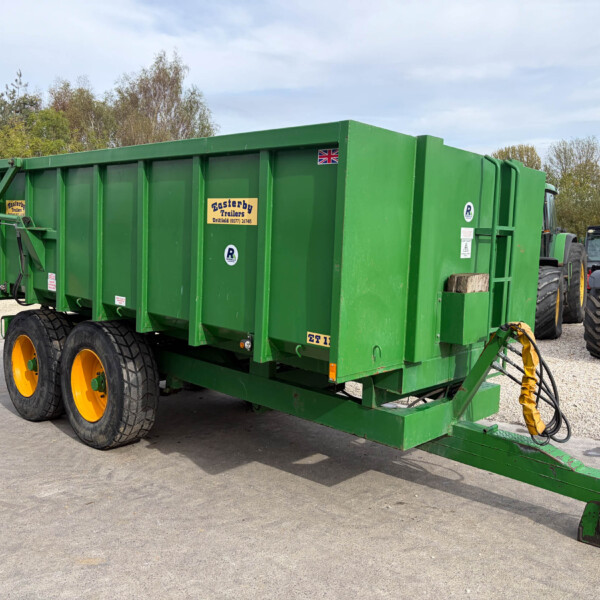 Green farm trailer with large yellow wheels, hitched to a tractor, parked on a gravel lot with trees in the background.