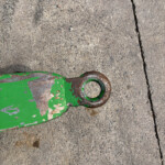 Close-up of a weathered green metal piece with a circular ring, resting on rough concrete pavement with peeling paint and rust.