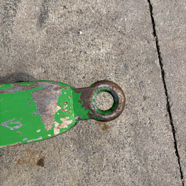 Close-up of a weathered green metal piece with a circular ring, resting on rough concrete pavement with peeling paint and rust.