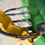 Close-up of hydraulic hoses connected to green agricultural equipment, with a yellow glove in the foreground and metal fittings visible.