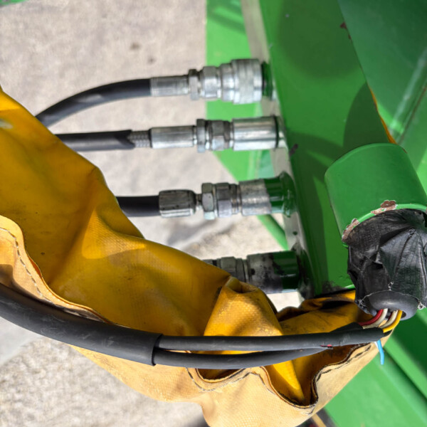 Close-up of hydraulic hoses connected to green agricultural equipment, with a yellow glove in the foreground and metal fittings visible.