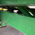 Underside view of green farm machinery frame with rust spots and angled metal supports, wheel and hydraulic components visible at lower left.