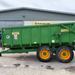 Green agricultural trailer with yellow wheels in a dealership lot, New Holland sign visible on the building behind it.