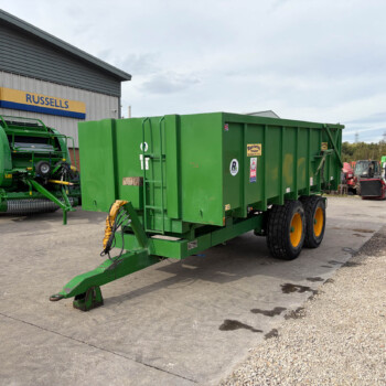 Large green agricultural trailer with a long hitch in a paved yard beside a workshop signboard.