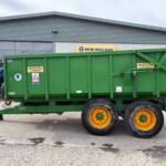 Green farm trailer with yellow wheels in a parking lot, New Holland branding visible on the building behind it