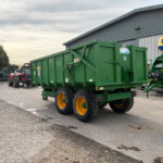 Green agricultural trailer with yellow wheels parked outside a farm equipment shop, New Holland Agriculture sign in the background.