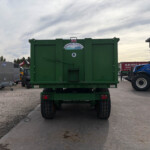Rear view of a large green agricultural trailer in a lot with other machinery and a blue tractor nearby. A UK flag sticker is on the trailer top center.