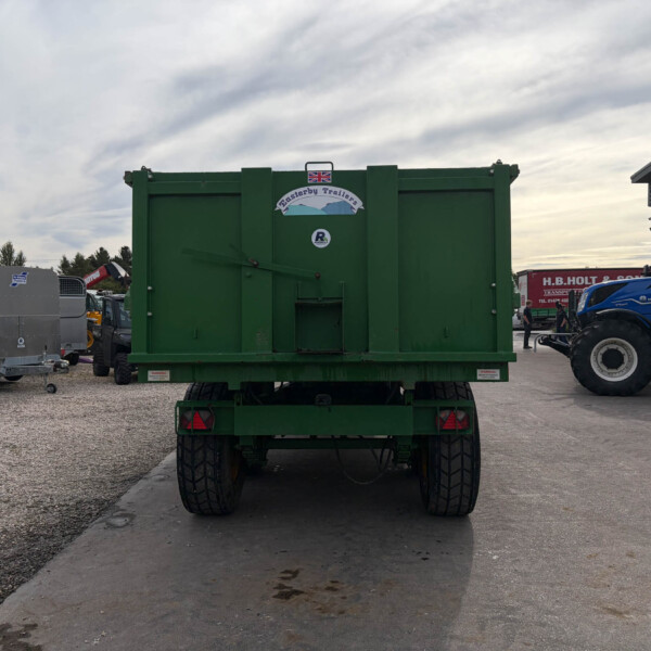 Rear view of a large green agricultural trailer in a lot with other machinery and a blue tractor nearby. A UK flag sticker is on the trailer top center.