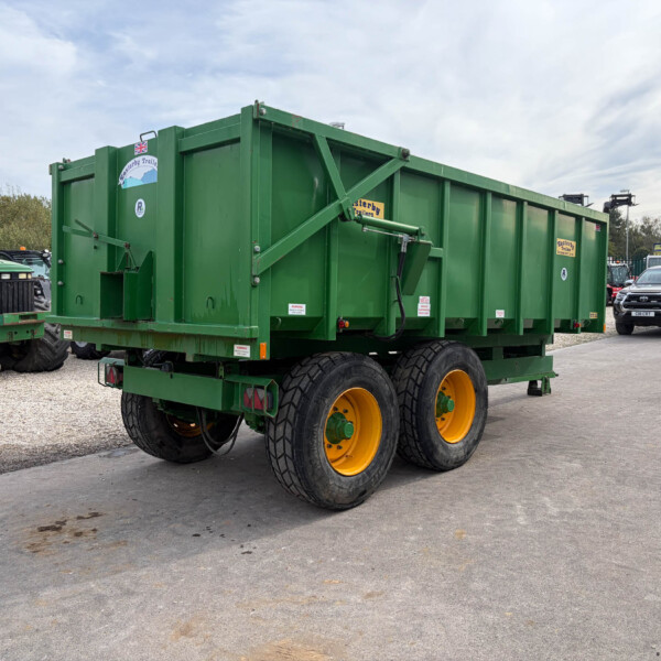 Green dump trailer with yellow wheels parked on a gravel lot beside a green tractor.
