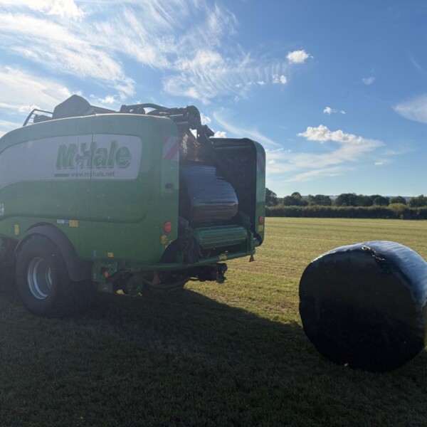 Green McHale baler parked in a grassy field with a large wrapped round bale nearby under a blue, sunny sky