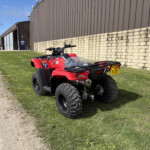Red all-terrain vehicle parked on a grassy area beside a stone wall and brown industrial building under a blue sky.
