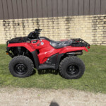 Red Honda ATV parked on grass with a beige stone wall and brown metal fence in the background.