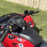 Close-up of red ATV handlebars and dashboard with safety stickers, parked on grass beside a stone wall.