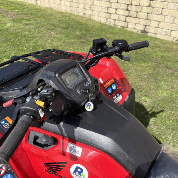 Close-up of red ATV handlebars and dashboard with safety stickers, parked on grass beside a stone wall.