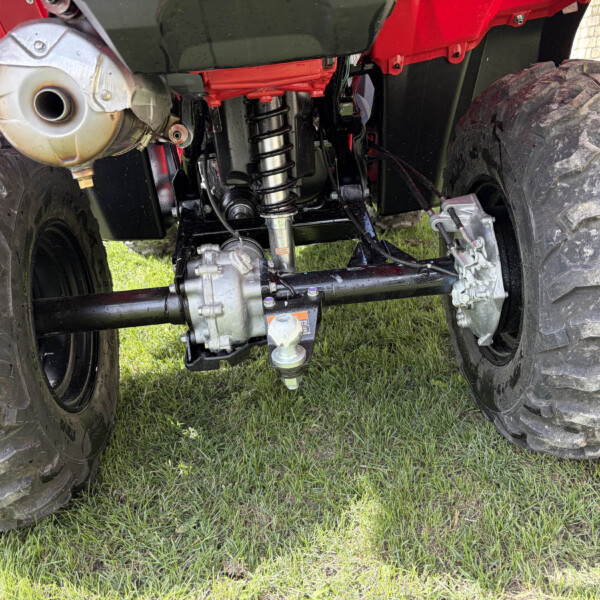 Underneath view of a red off-road vehicle showing suspension, drive shaft, and large muddy tires on grass.