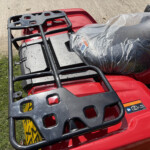 Close-up of a red motorcycle with a black metal luggage rack and a gray plastic-covered seat/bag strapped on top.