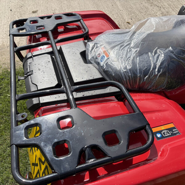 Close-up of a red motorcycle with a black metal luggage rack and a gray plastic-covered seat/bag strapped on top.