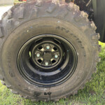 Close-up of a large off-road tire with mud on the tread, mounted on a black rim, resting on green grass.