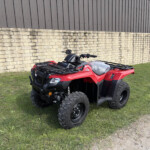 Red ATV parked on a grassy area beside a beige stone wall and gray metal-clad building in the background.