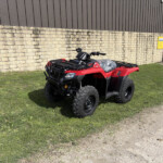 Red ATV parked on grass beside a stone wall and brown metal building exterior with a yellow sign in the background.
