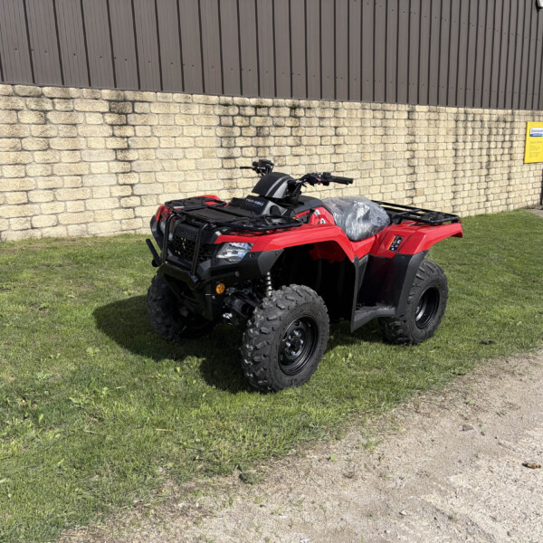 Red ATV parked on grass beside a stone wall and brown metal building exterior with a yellow sign in the background.
