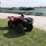 Red Honda ATV parked on grassy area with industrial buildings in the background and a person in protective white suit on the left.