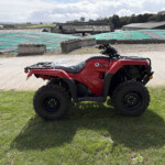 Red Honda ATV parked on grass with industrial buildings and a tarped area in the background.