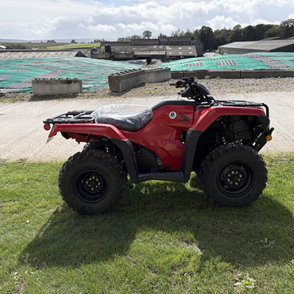 Red Honda ATV parked on grass with industrial buildings and a tarped area in the background.
