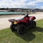 Red ATV parked on grass near a gravel area with a construction yard and warehouses in the background.