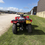 Red off-road ATV parked on a grassy area near industrial buildings and a gravel yard with blue sky above.