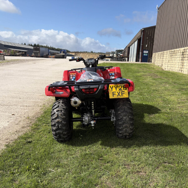 Red off-road ATV parked on a grassy area near industrial buildings and a gravel yard with blue sky above.