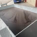 Metal flatbed trailer with diamond-plate sides and a wet, textured deck in an outdoor industrial area.
