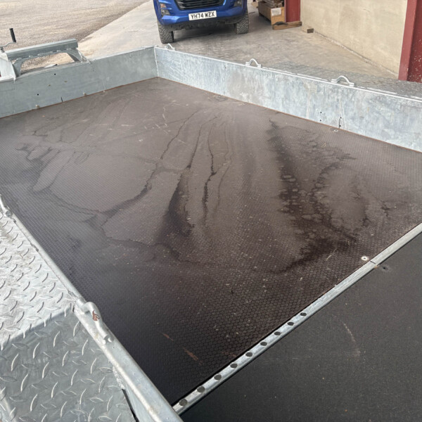 Metal flatbed trailer with diamond-plate sides and a wet, textured deck in an outdoor industrial area.