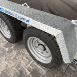 Diamond-plate trailer bed with dual-axle wheels and tires visible, showing a sturdy metal frame and wheel hubs. Note: blue lettering on the side indicates the trailer brand.