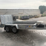Industrial outdoor loading area with a metal flatbed trailer and perforated ramp sections beside a tall gray concrete wall under an overcast sky.