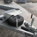Metal utility trailer with a mounted spare tire and fold-down ramp in an outdoor yard.