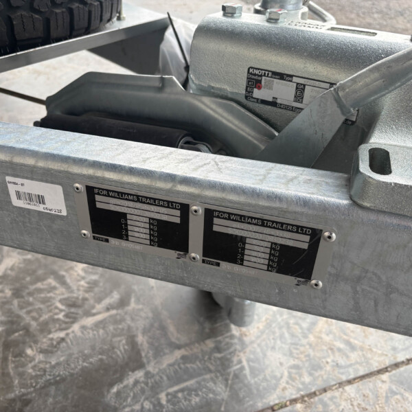 Close-up of a metal trailer hitch/frame showing data/weight rating plates and bolts, with a spare tire in the background on a rack.