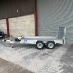 Blue Isuzu pickup parked beside a large metal warehouse with corrugated doors and a silver trailer in a construction yard.