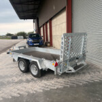 Blue Isuzu pickup parked beside a silver utility trailer in an industrial loading yard by a corrugated metal warehouse door under a cloudy sky.