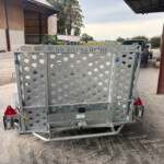 Rear view of a perforated metal trailer gate in a concrete lot, with a blue truck and farm buildings in the background