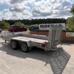 Metal trailer parked in a lot with cars and a large white Russell sign in the background, clear sky above.