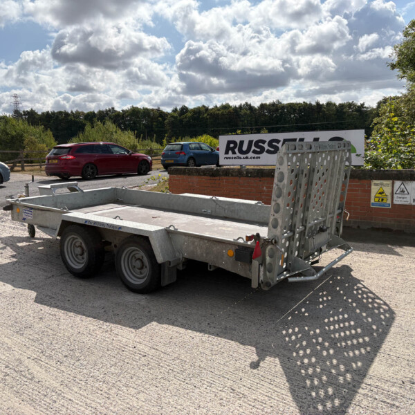 Metal trailer parked in a lot with cars and a large white Russell sign in the background, clear sky above.