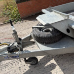 Trailer frame with a mounted spare tire, metal ramp, and coiled cables on a dirt surface beside a brick wall, likely ready for towing.