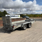 Vehicle trailer with metal side rails in a paved lot, a brick wall, and a large Russell's Trailers sign in the background under a cloudy sky