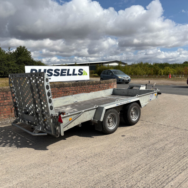 Vehicle trailer with metal side rails in a paved lot, a brick wall, and a large Russell's Trailers sign in the background under a cloudy sky