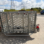 Front view of a perforated metal trailer with circular holes on a sunny roadside; a blue car is visible in the distance.