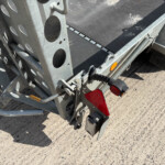 Close-up of a metal utility trailer rear corner showing perforated beams, a coiled suspension spring, and red taillight lenses on a gravel surface.